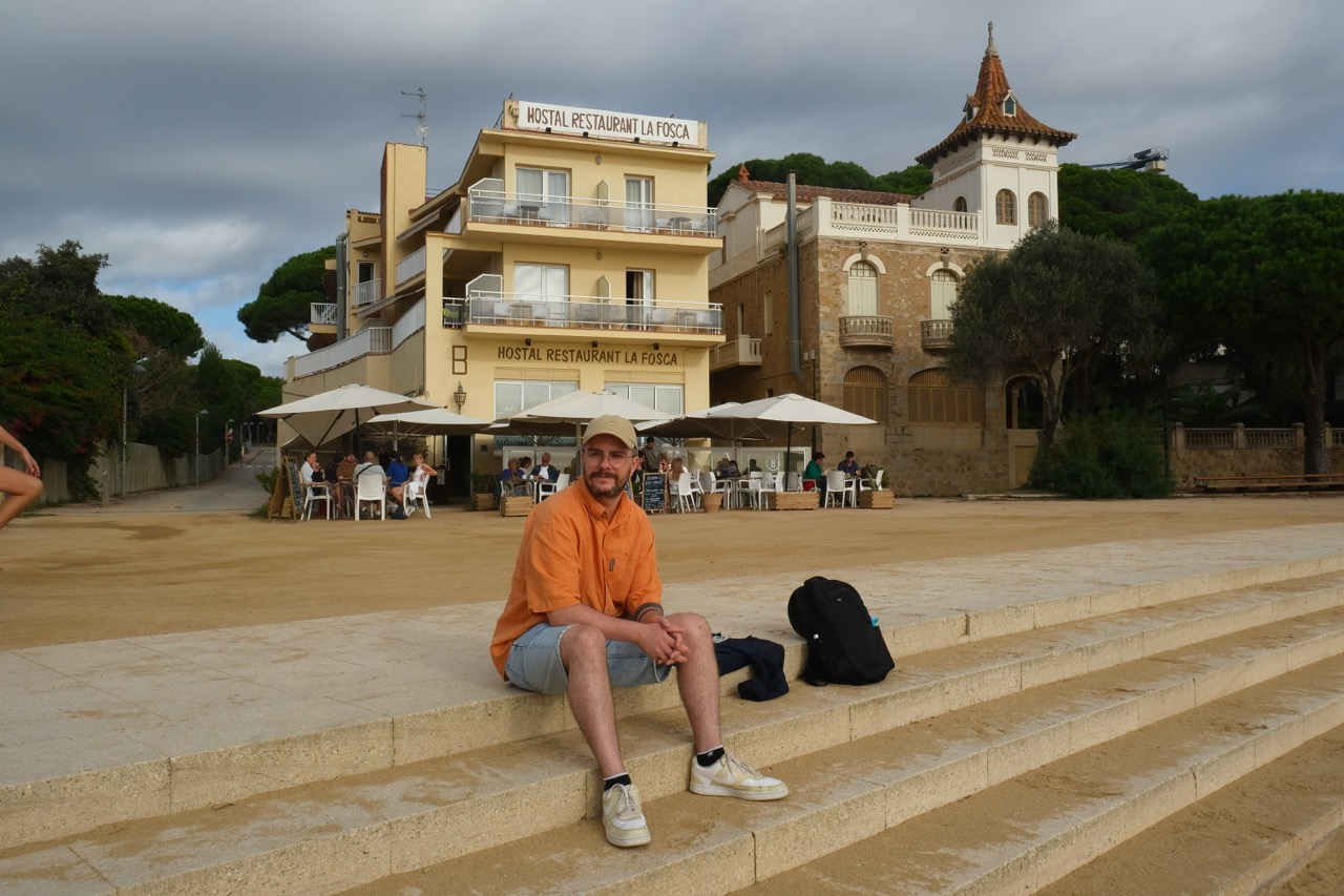 Rob sitting on stone steps at La Fosca beach with Mediterranean architecture behind